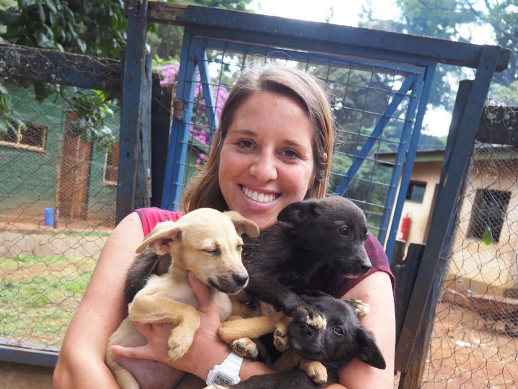 Young woman holding three puppies, two are black, one is golden. They're outside on the grounds of a dog shelter